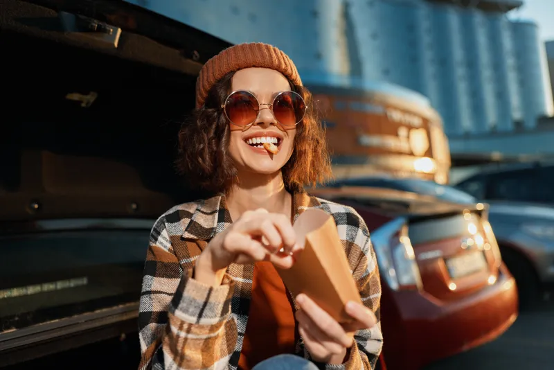 Woman car fries smile sunglasses beanie trunk golden hour candid lifestyle: young woman eating takeaway fries by an open car trunk at sunset, authentic emotional storytelling with warm mindful living