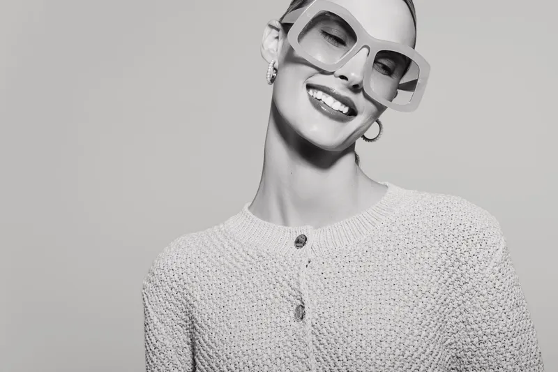 Smiling caucasian woman with fair skin wearing oversized sunglasses and a knitted cardigan poses against plain background. Concept of fashion, style, and confident female model portrait.