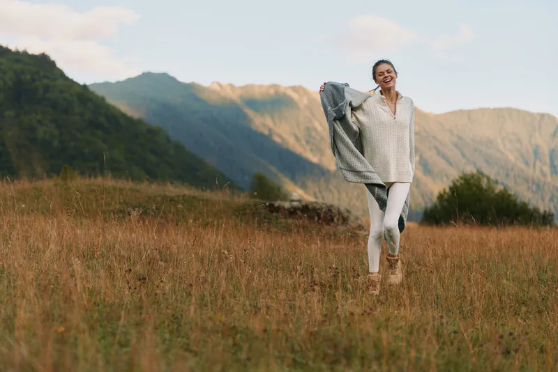 Joyful woman outdoors in a sunlit field, wearing casual white outfit, breeze lifting her hair, mountains in the background, relaxed lifestyle moment, natural colors and gentle breeze in air
