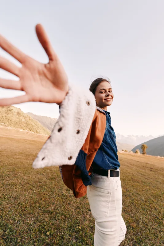 Woman smiling with backpack in mountains field reaching hand toward camera. Young traveler in denim shirt and white pants, outdoor adventure and hiking portrait full of freedom.