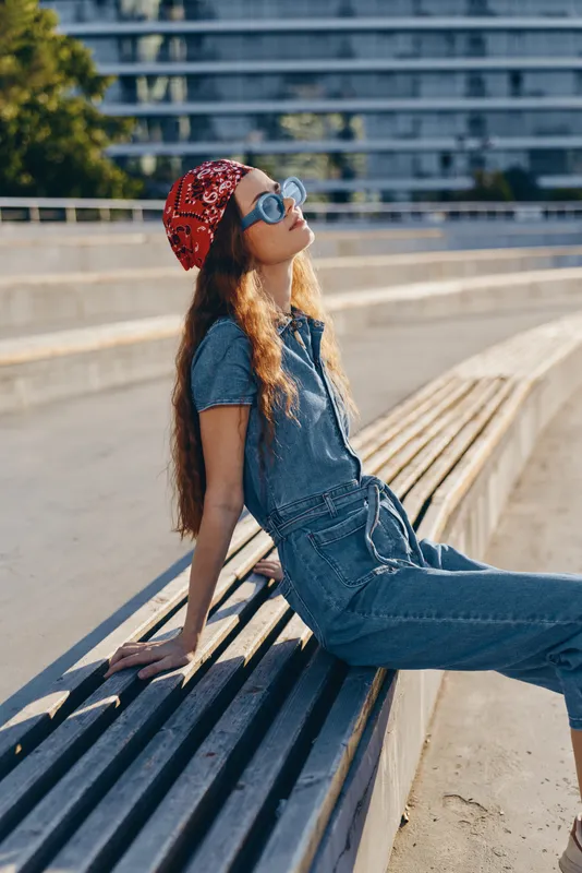 Relaxed young woman in denim jumpsuit and red bandana sitting on urban wooden bench enjoying sunny day outdoors with sunglasses and casual style
