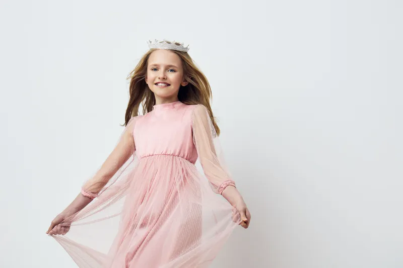 Joyful girl in a flowing pink dress with a crown smiling against a clean white background, capturing the essence of childhood innocence and elegance