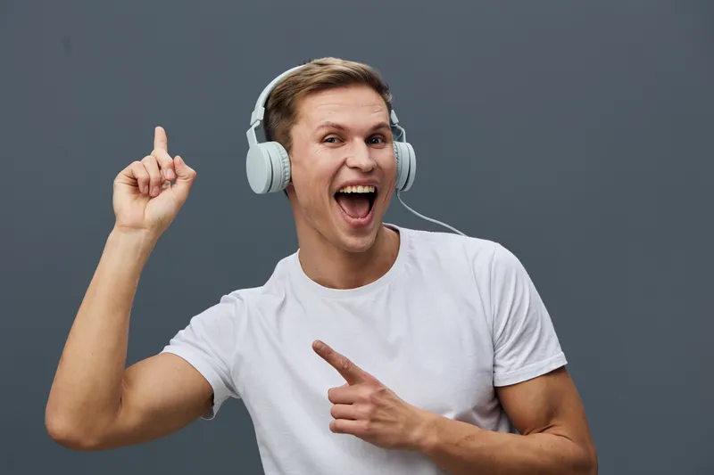 happy young man wearing white t-shirt and headphones listening to music pointing finger showing excitement isolated on dark gray background digital content generation concept