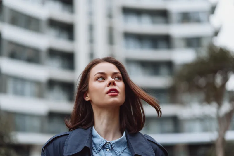 A young woman with brunette hair walks along a city street in a blue denim shirt, looking up with confidence, wind in her hair, and a modern architectural backdrop.