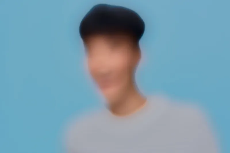 Blurred man wearing casual gray shirt and black cap on soft blue background, portrait with indistinct facial features creating mysterious and artistic mood.
