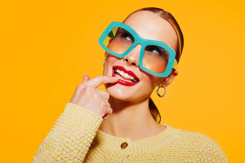 Young woman with large blue sunglasses and red lipstick smiles thoughtfully, wearing a yellow sweater against a bright orange background. Concept of fashion, style, and vibrant colors.