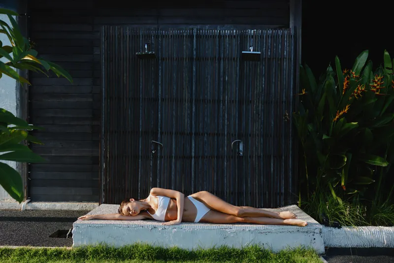 Relaxed woman in a white swimsuit lying beside an outdoor shower in a tropical garden, sunlit scene, capturing a serene lifestyle, wellness, and simple self care mood