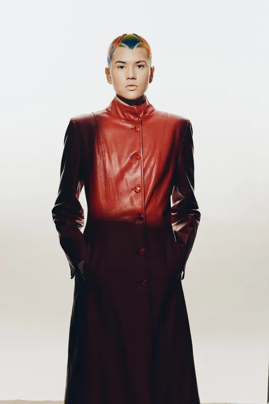 woman wearing long red leather coat with colorful braided hair, standing against light background, serious expression, modern fashion style