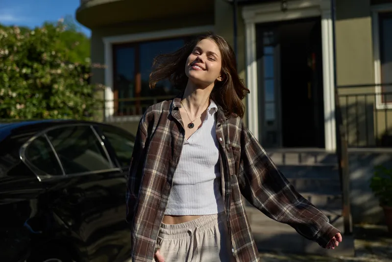 Woman lifestyle portrait outdoors in sunny weather, wearing plaid jacket over light blouse, relaxed pose, candid smile, cheerful mood, modern casual fashion, suburban backdrop