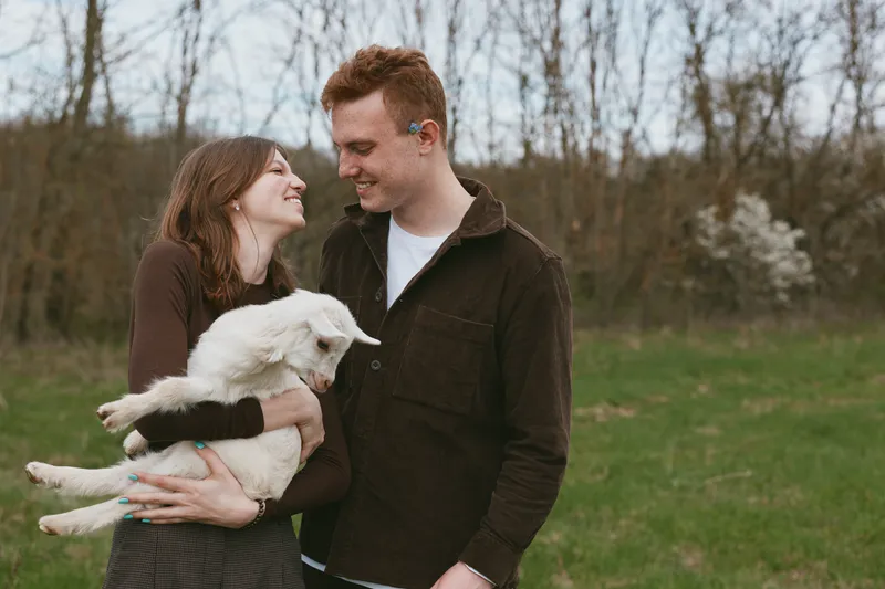 Young couple embraces outdoors while the woman holds a white baby goat, sharing a joyful moment in casual autumn clothing with bare trees in the background.