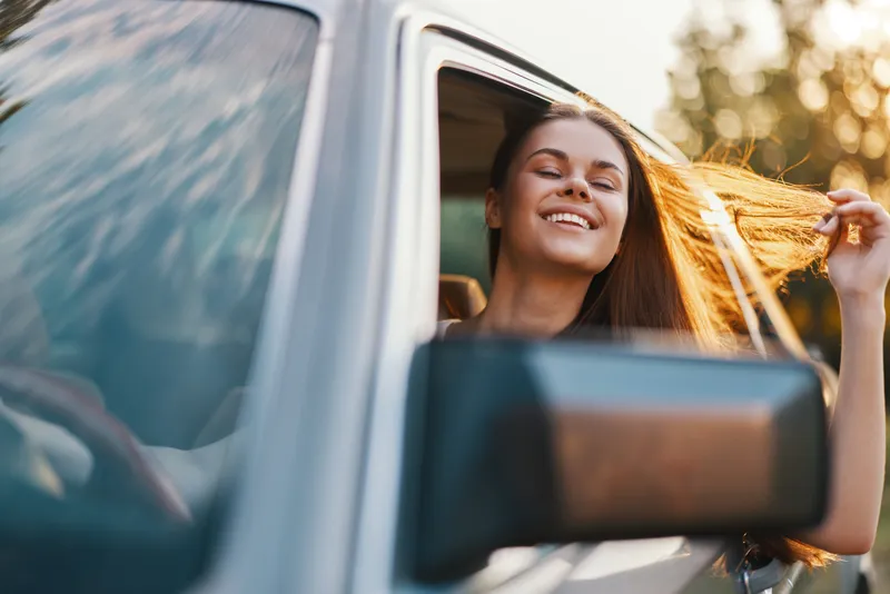 Young woman in a car enjoying a sunny day, with long hair flowing in the breeze, expressing joy and freedom, perfect for seasonal travel themes.
