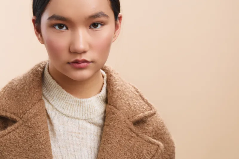 Asian woman wearing a cozy sweater and coat looks directly at the camera, presenting a poised, calm portrait with a soft, warm mood and contemporary style.