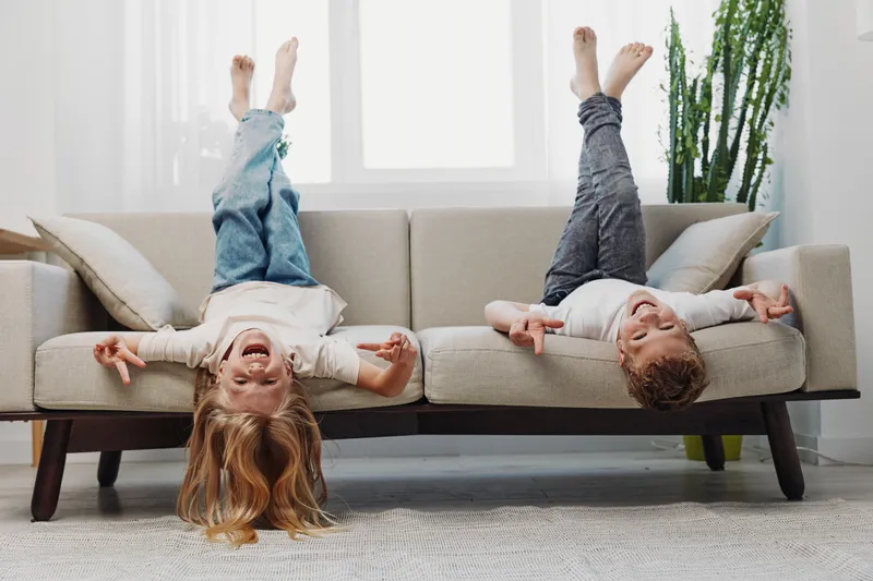 Two happy children lying upside down on a beige sofa with legs raised, laughing and having fun in a bright living room with a large window and indoor plant.