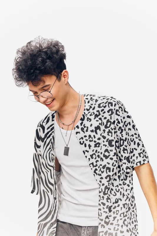 young man with curly hair smiling cheerfully in trendy mixed animal print shirt and glasses, wearing layered necklaces against white background