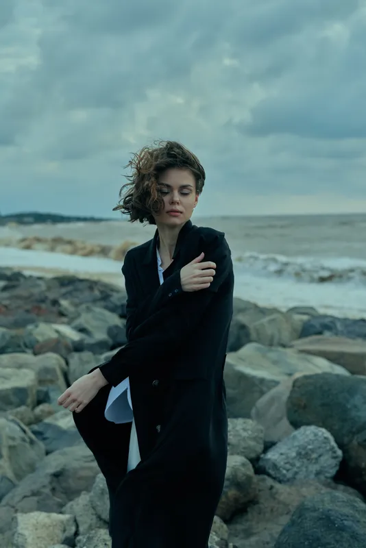 Windy seaside fashion, woman in a dark coat on a rocky shore, outdoor mood with coastal breeze and moody clouds, dramatic silhouette against rugged stones, gray water, minimal editorial vibe