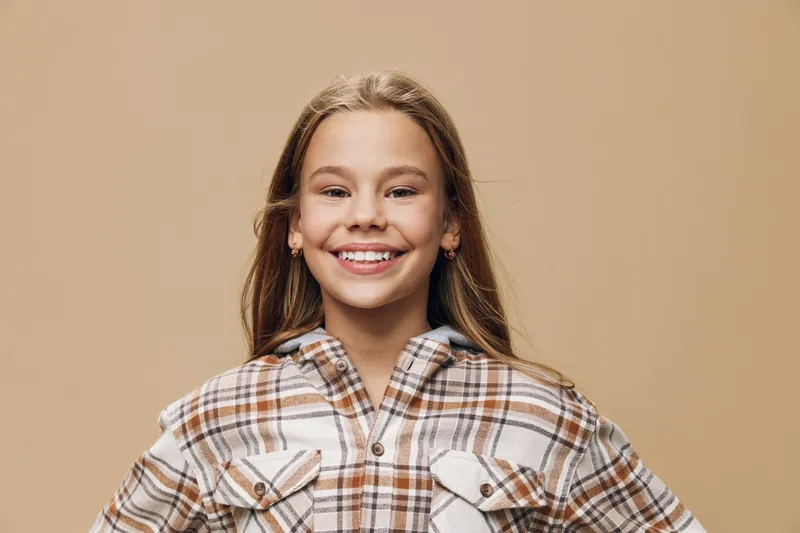 Girl smiling and standing with hands on hips wearing plaid shirt with beige background. Teenager confident happy and casual in light brown and white checkered attire.