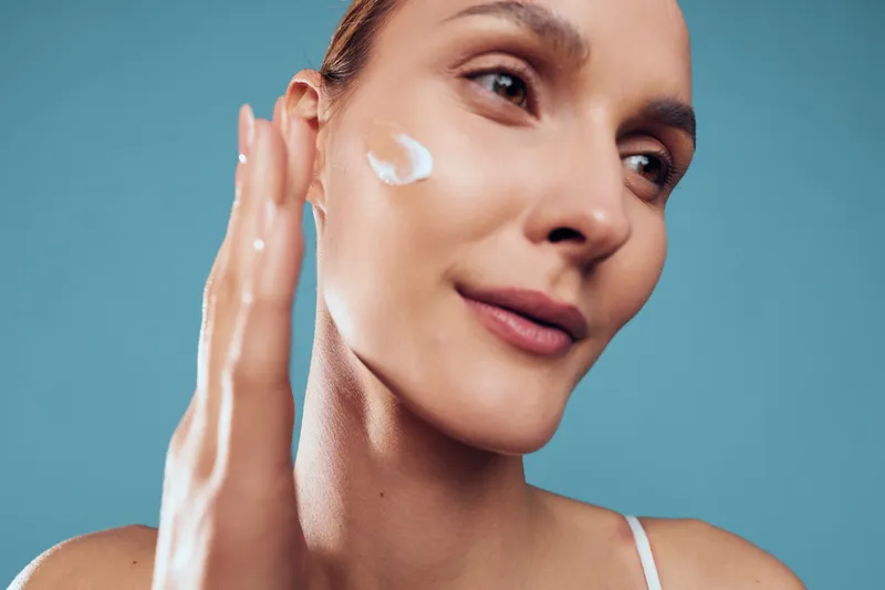 Young woman applying facial cream on clean skin, close up portrait, natural makeup, soft light, blue background, skincare routine, healthy glowing complexion, beauty care