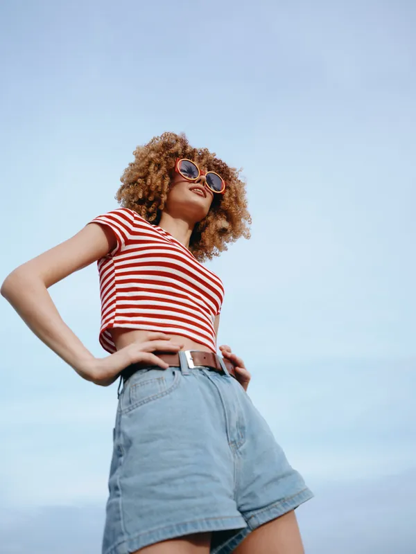 Young woman with curly hair wears a red striped crop top and high waist denim shorts, standing with hands on hips under a clear blue sky, exuding confidence and style.