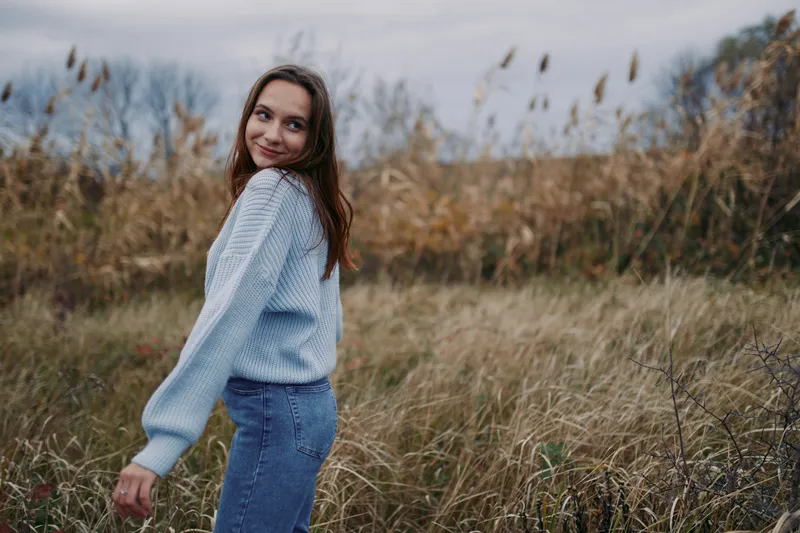 Candid portrait of a young woman in a cozy sweater walking through tall grass, looking back with a genuine smile, natural light and honest framing emphasizing authentic emotion.