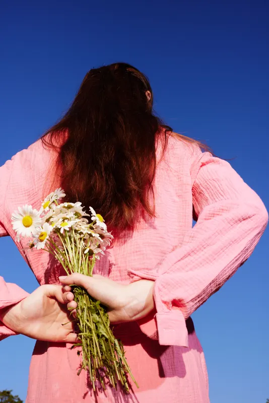 Young woman holding flowers behind her back in a pink dress, standing against a clear blue sky, conveying a cheerful and playful emotion