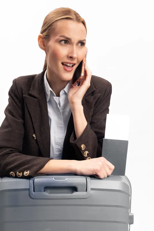 Young woman in brown blazer talks on phone while holding passport and leaning on gray suitcase. Concept of travel, business trip, communication and modern lifestyle.