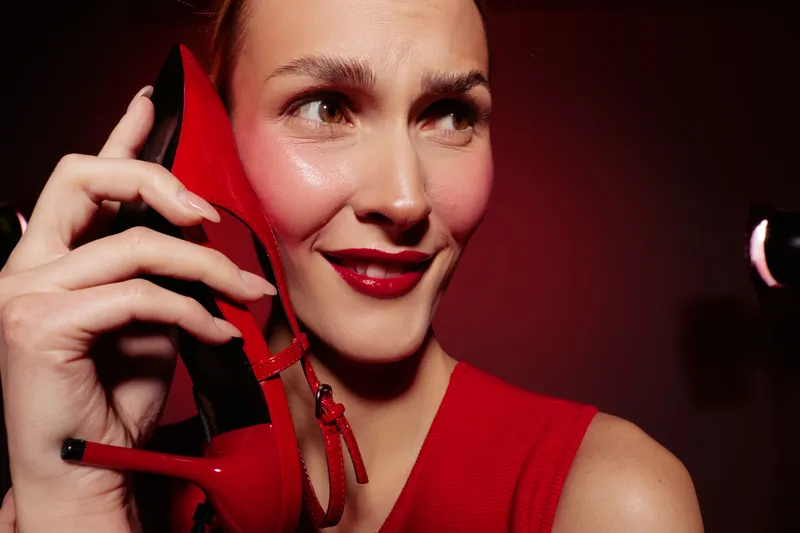 Smiling woman in red dress holds red high heel shoe near face, showing playful expression. Concept of fashion, style, beauty, and confidence in studio lighting.
