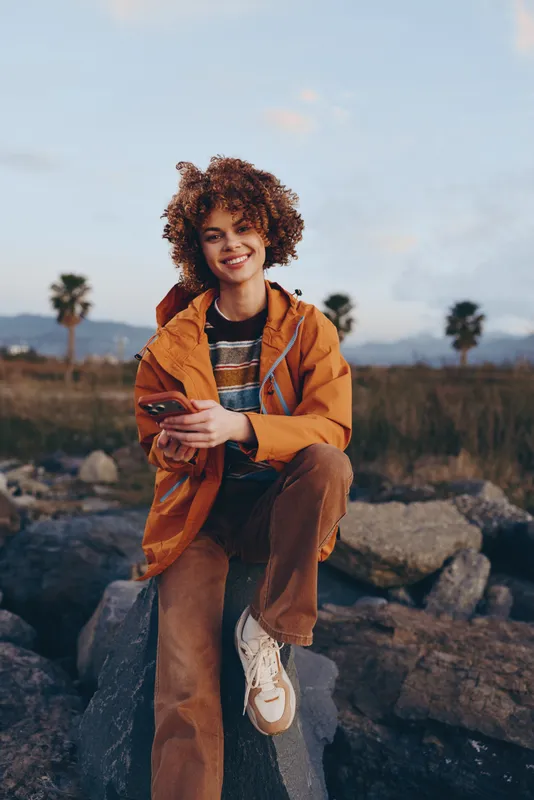 Smiling woman outdoors, curly hair, orange jacket, relaxed lifestyle pose on rocky ground, warm evening light, scenic horizon, shallow depth of field