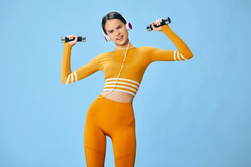 Fit woman in orange workout outfit lifts small dumbbells while wearing headphones, smiling confidently in a light blue studio setting; fitness, sport and active lifestyle concept.