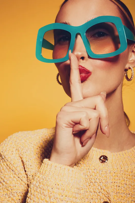 Stylish woman in large blue glasses and yellow sweater makes silence gesture with finger on lips. Bright yellow background highlights fashion and secrecy concept in studio portrait.