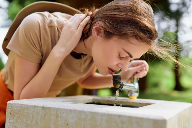 Woman drinking water from a public fountain in a sunny park, young casual female leaning to sip fresh outdoor water, summer greenery and natural hydration moment.