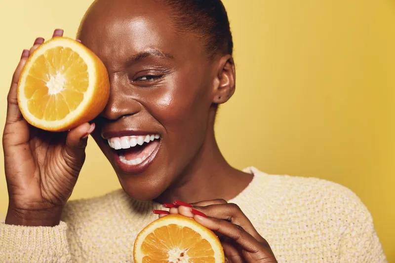 Smiling dark skinned African woman holds two halves of fresh orange fruit near face on yellow background. Concept of healthy lifestyle, vitamin, skincare and natural beauty.