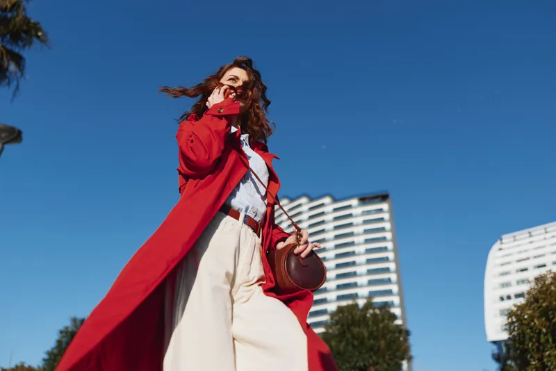 A stylish woman in a red coat walks outdoors in an urban setting, sunlight bright, wind blowing her hair. A bold fashion moment highlighting confidence, street style, and contemporary city living.