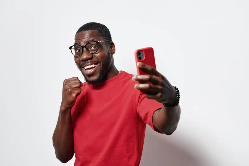 African American man with dark skin wearing glasses and red shirt taking selfie with smartphone smiling and showing fist gesture on white background.