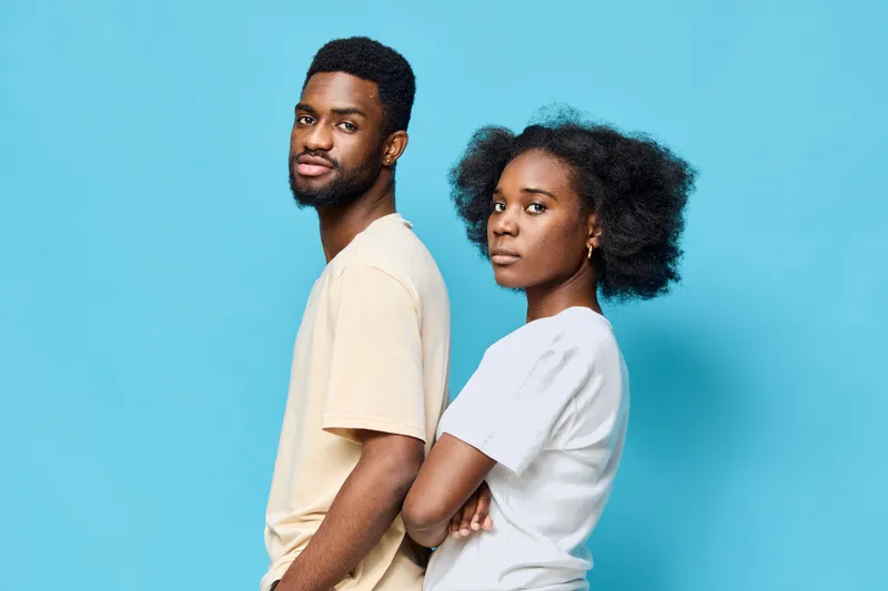 A confident young Black couple in casual t shirts stands back to back against a bright blue background, embodying strength and unity in a contemporary style