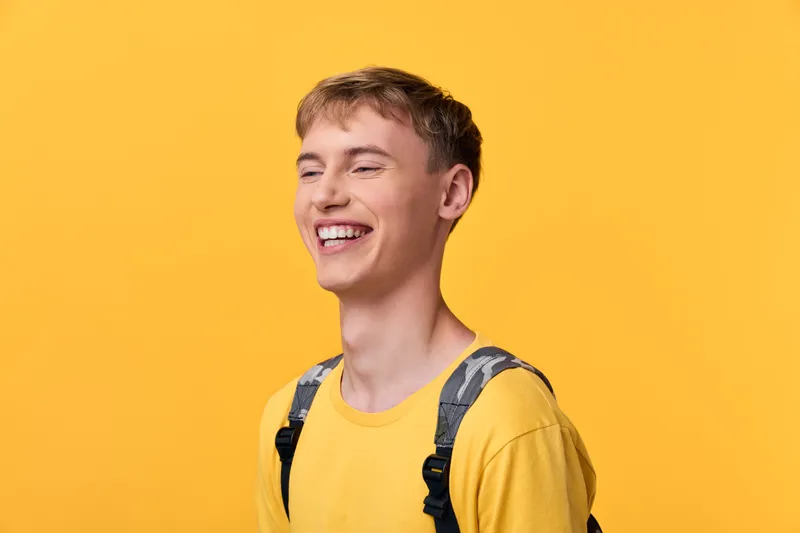Young man in a yellow shirt with a backpack smiles, bright studio shot conveying cheerful mood. Casual lifestyle imagery for youth, education, or brand storytelling with positive energy.