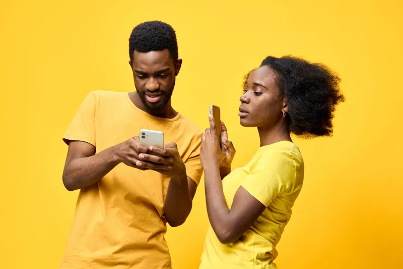 Young African American couple interacting with smartphones against a vibrant yellow background, expressing curiosity and playfulness in bright matching outfits