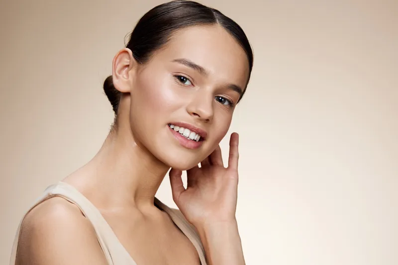 Portrait of a smiling young woman with natural beauty, wearing minimal makeup and a beige top, set against a soft neutral background for a fresh, glowing look