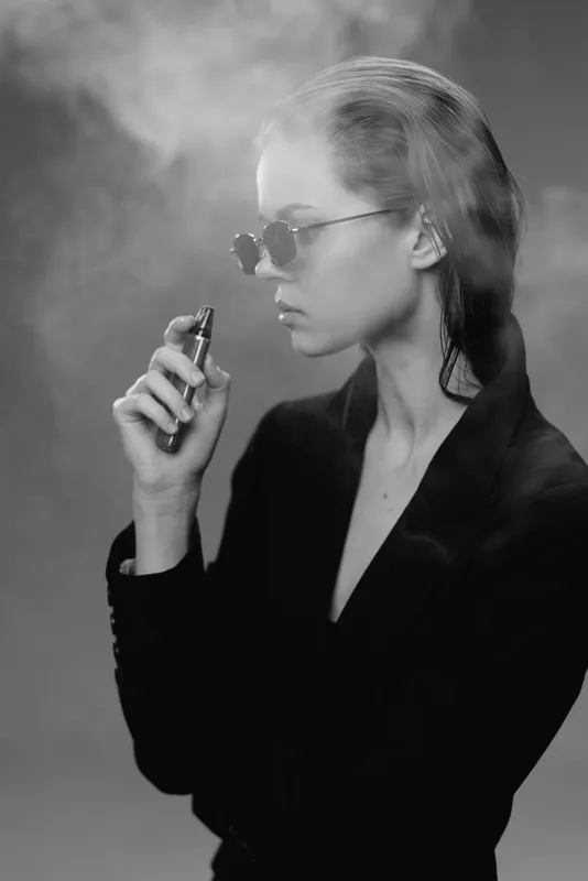 Black and white portrait of a serious woman vaping with smoke around her in stylish sunglasses and dark clothing.