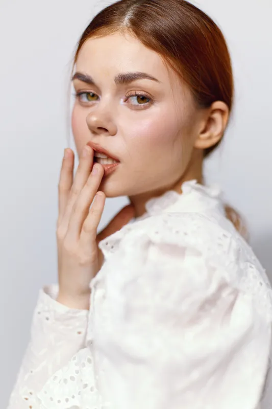 Young woman with brown hair touching her lips, wearing a white blouse, expressing a thoughtful emotion against a light background, perfect for beauty and fashion themes