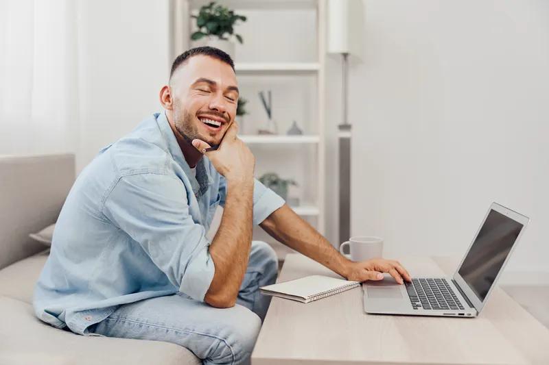A cheerful man working on a laptop at home, thoughtfully smiling and engaged in productivity. Bright, airy living space conveys a modern lifestyle.