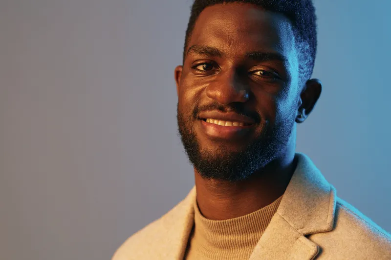 Confident Black man smiling in a stylish outfit with a soft blue background, showcasing a modern and relaxed concept Description includes fashion, emotions, and colors