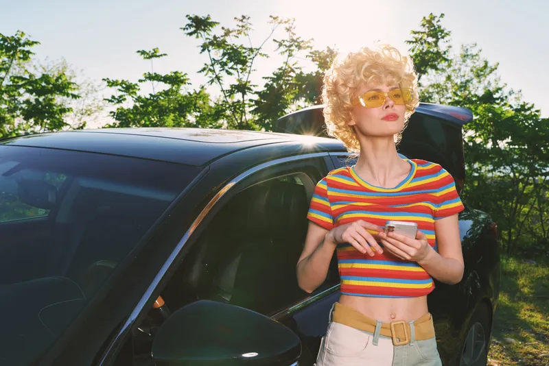 Woman outdoors, retro fashion, striped crop top, standing beside a car in bright sunlight, casual lifestyle, vintage colors, lush green background, carefree moment
