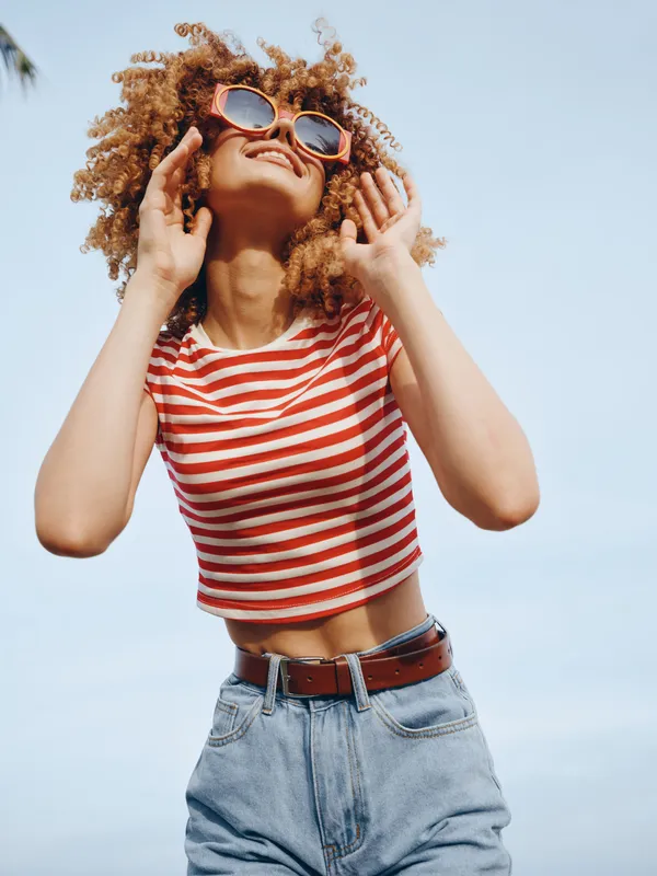Vibrant portrait of a joyful woman in a red and white striped crop top and denim jeans, captured outdoors under a bright sky, exuding carefree summer energy.