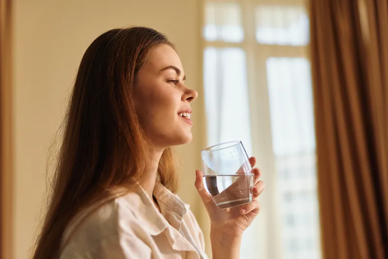 Woman drinks water from a glass with a gentle smile in a bright home morning portrait by a window, natural light creating a relaxed, healthy daily routine and calm mood.