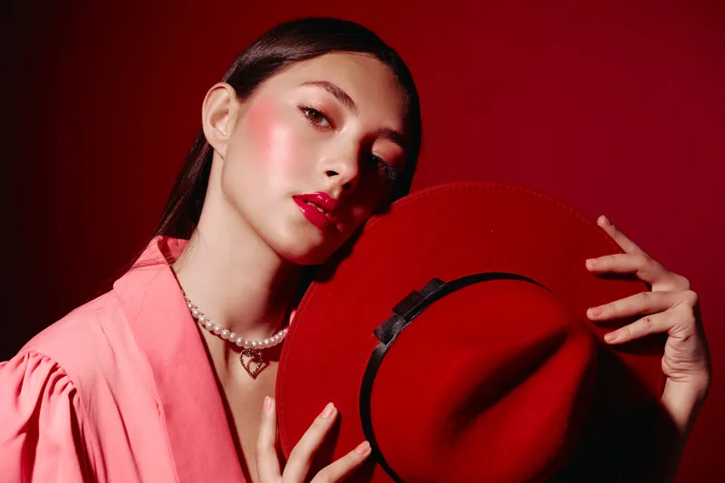 Young woman with glossy red lipstick and blush poses holding a red hat in front of a red background. Styled in pink jacket and pearl necklace. Creative fashion and beauty concept.