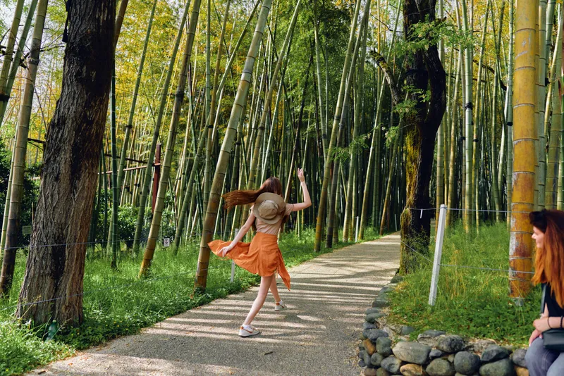 Woman in a hat and orange dress walking and twirl on a gravel path through a bamboo forest in summer nature, joyful movement and carefree exploration under sunlight and leafy trees.