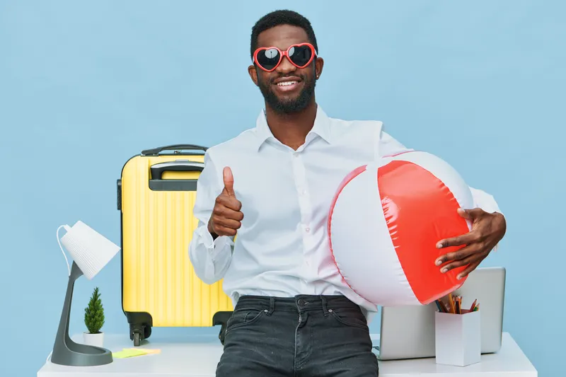 Joyful man with a beard and sunglasses smiling and holding a beach ball, standing in front of travel accessories including a yellow suitcase, laptop, and small plant, on a light blue background. Summe