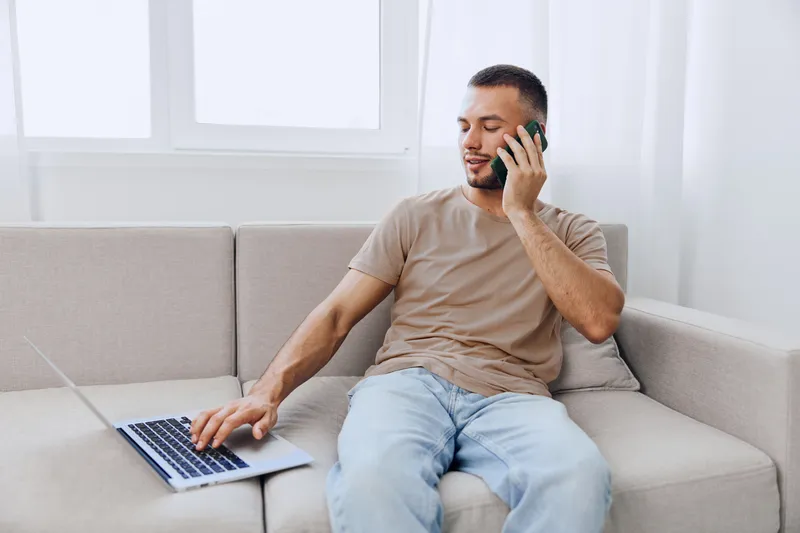 A focused man talking on the phone while using a laptop, exuding casual elegance in a modern living room with soft lighting and neutral colors.
