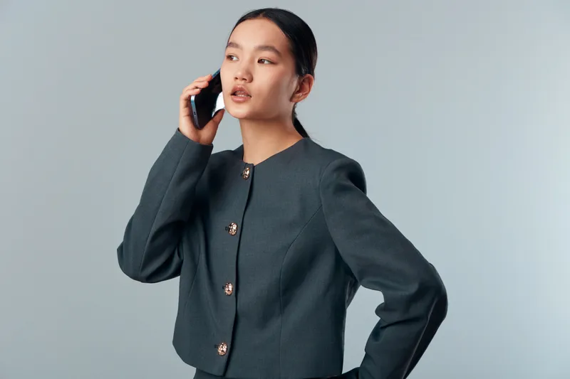 Woman talking on phone with hand on hip in gray jacket against neutral background. Young female model with serious expression making call, communication and technology in modern lifestyle concept.