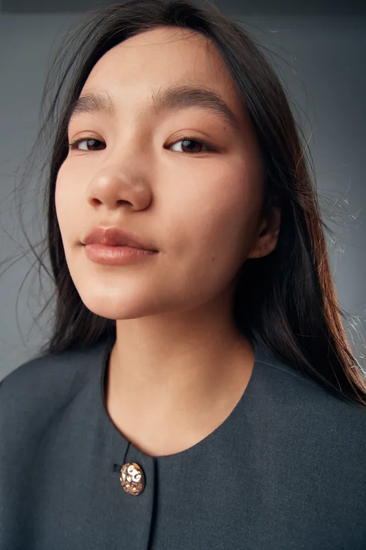 Portrait of young woman with long dark hair and natural makeup looking at camera against gray background wearing dark clothing with single decorative button closeup for beauty or fashion concept.